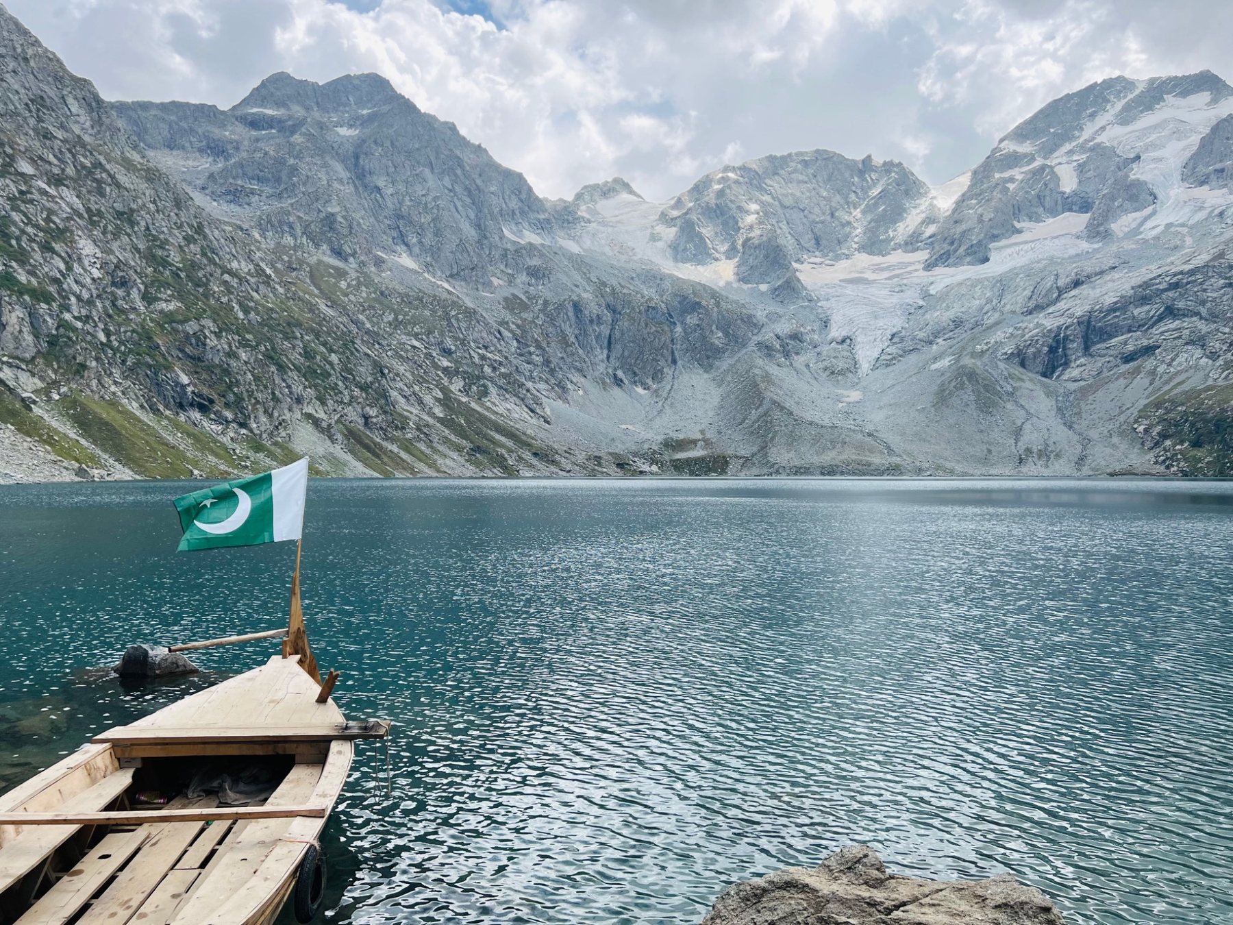A wooden boat with a Pakistani flag on a turquoise glacier lake below snow peaks